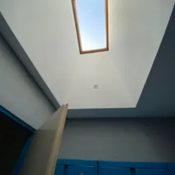 Pomieszczenie po malowaniu: biały sufit z oknem dachowym i szare ściany nad niebieskimi szafkami. Widoczna krawędź płyty gipsowej.