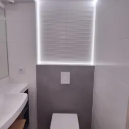 Nowoczesna toaleta z podświetleniem LED, szare płytki imitujące beton, biała ceramika i drewniane półki. Minimalistyczny design z dbałością o detale.