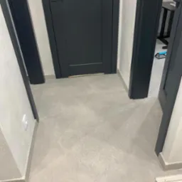 Szare płytki podłogowe w korytarzu, widoczne fragmenty ścian i ciemne futryny drzwi. Minimalistyczny design, neutralna kolorystyka. Ukończony projekt, czysto i schludnie.