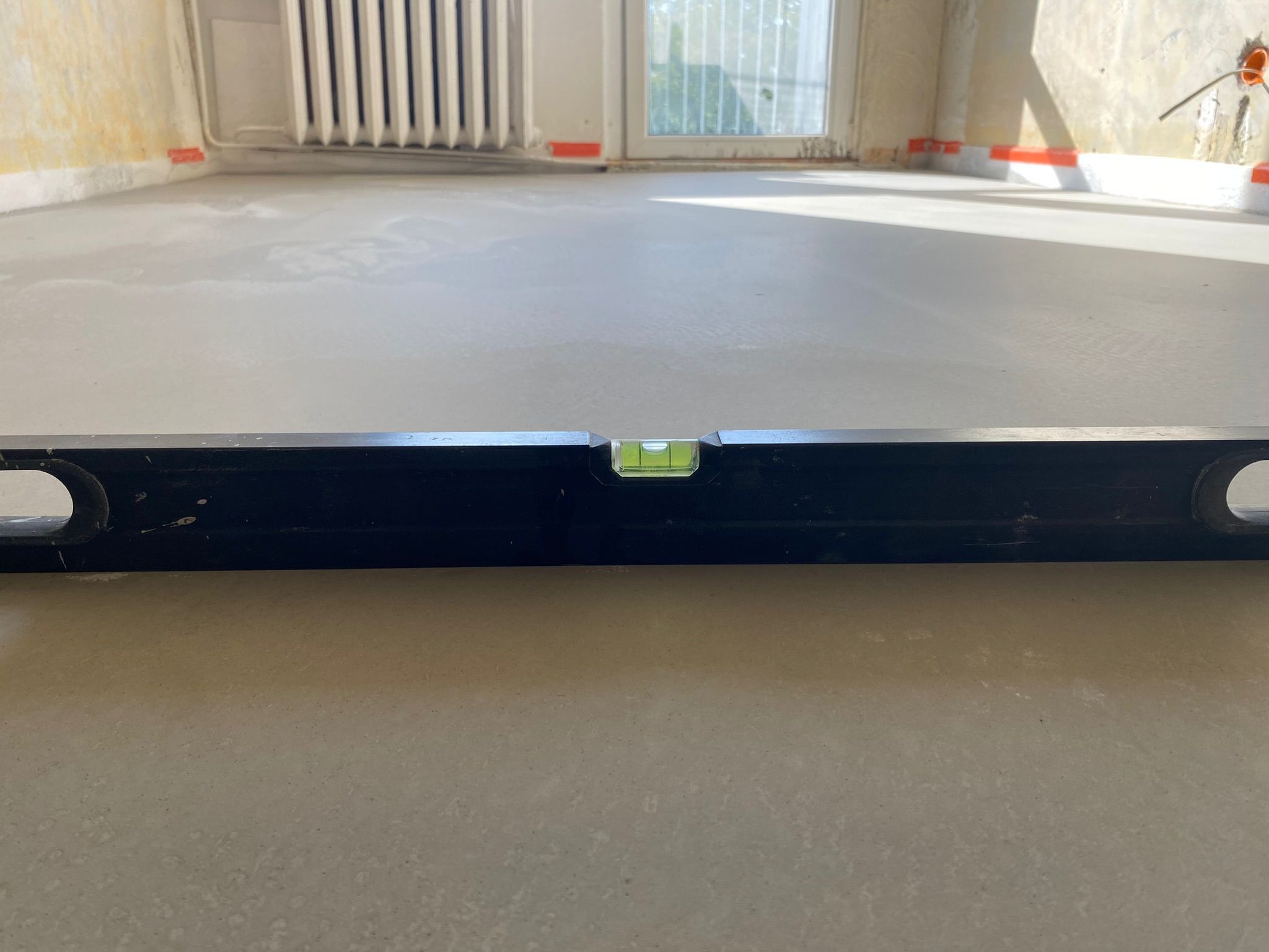 Wyrównana wylewka podłogowa sprawdzana poziomicą w remontowanym wnętrzu. Widoczne zabezpieczenia ścian taśmą. W tle okno i grzejnik.