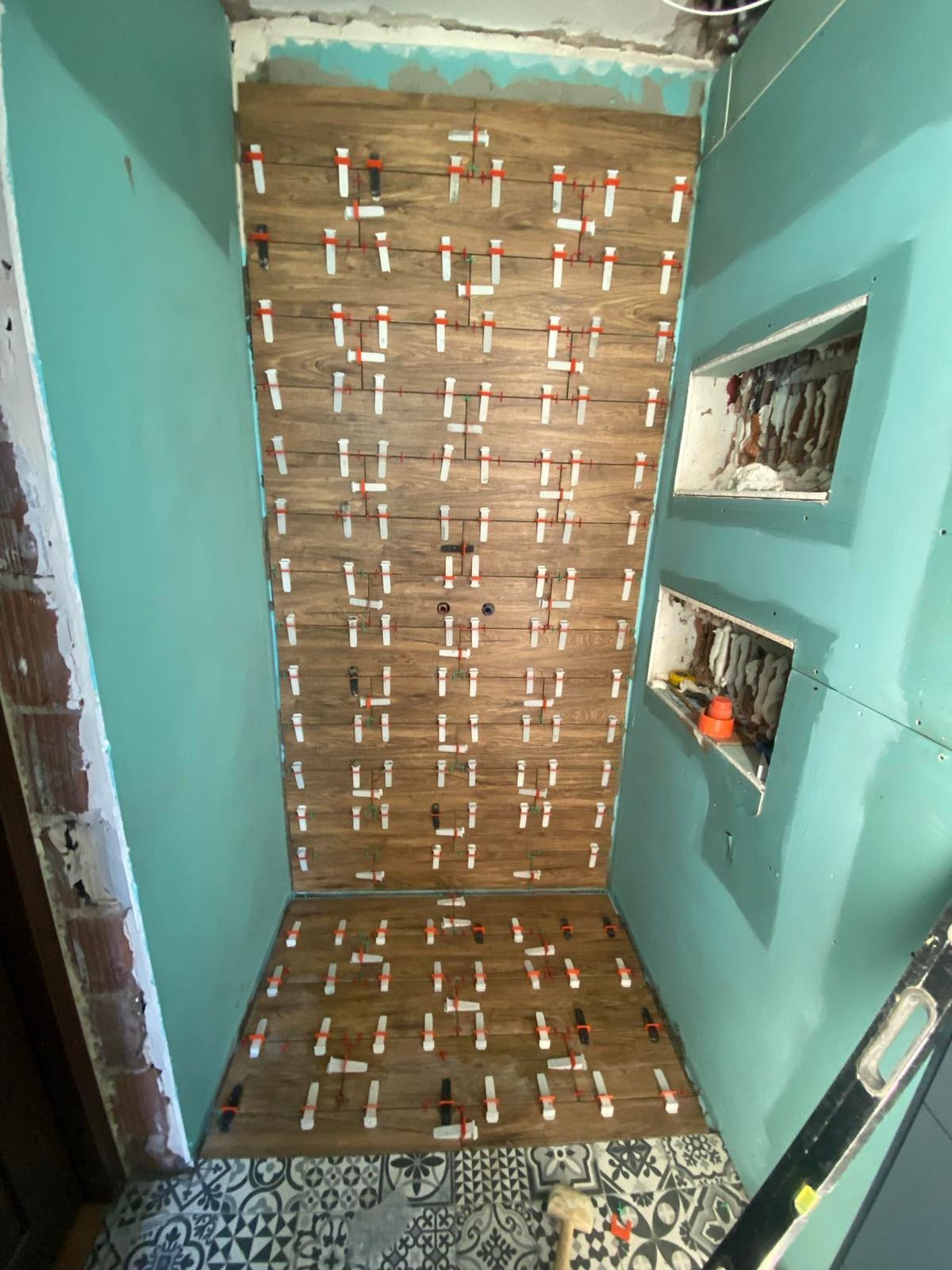 Układanie drewnopodobnych płytek w kabinie prysznicowej z półkami wnękowymi. Widoczne klipsy poziomujące. Na podłodze wzorzyste płytki.