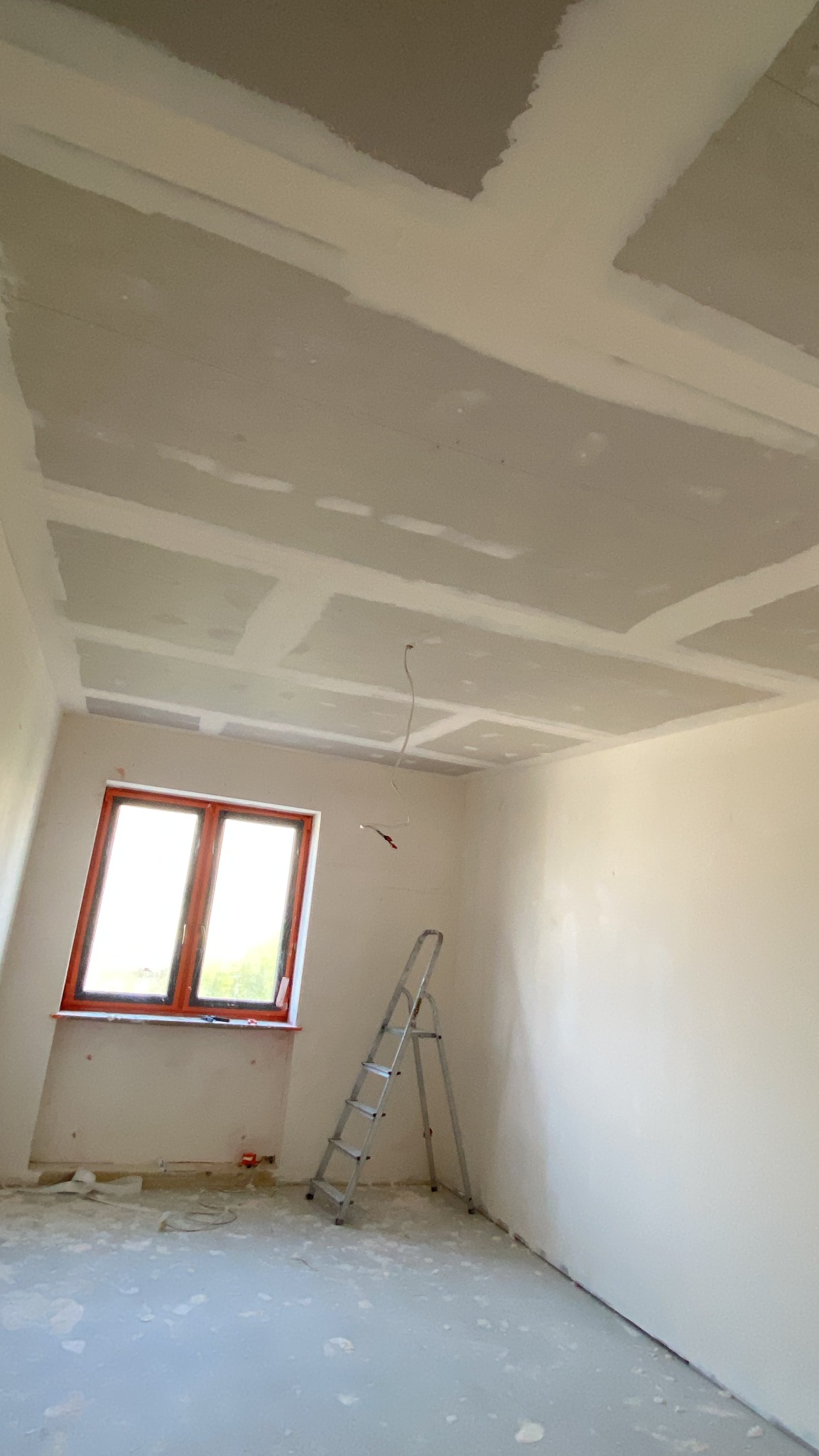 Pokój w trakcie prac wykończeniowych: zamontowane płyty karton-gips na suficie, drabina oparta o ścianę przy oknie. Widoczne niedoskonałości i ślady po pracach.