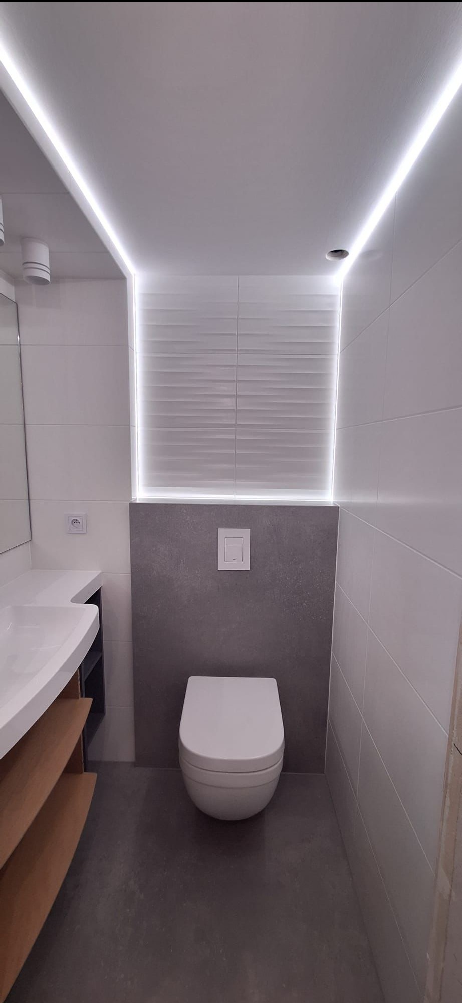 Nowoczesna toaleta z podświetleniem LED, szare płytki imitujące beton, biała ceramika i drewniane półki. Minimalistyczny design z dbałością o detale.