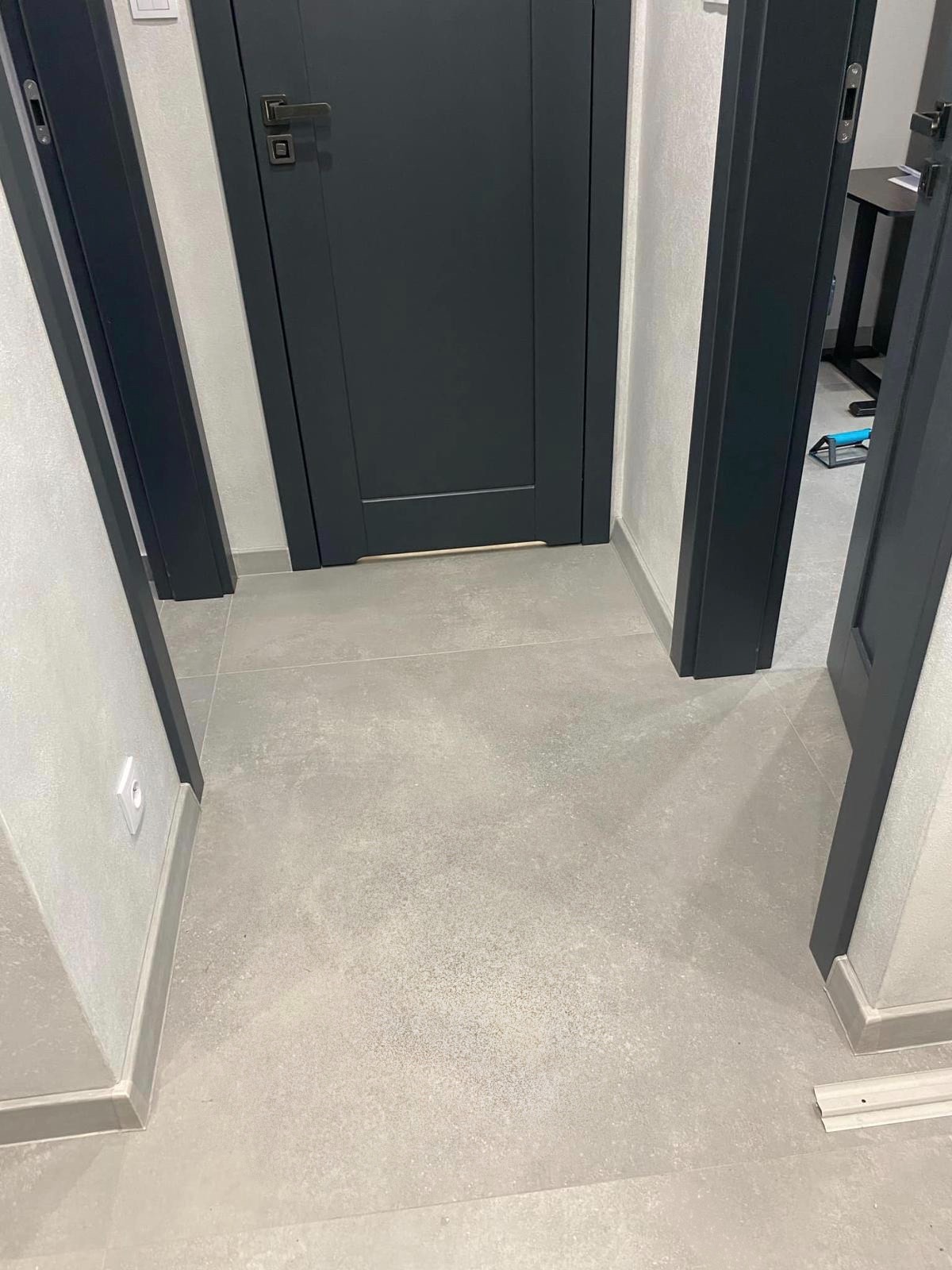 Szare płytki podłogowe w korytarzu, widoczne fragmenty ścian i ciemne futryny drzwi. Minimalistyczny design, neutralna kolorystyka. Ukończony projekt, czysto i schludnie.