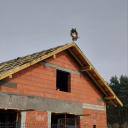 Usługi Ogólnobudowlane Specjalność Ciesielstwo - Maciej Hirsz - Budowa domu z czerwonej cegły z drewnianą konstrukcją dachu w trakcie realizacji. Na szczycie dachu wieniec świąteczny. Widok zza ogrodzenia.