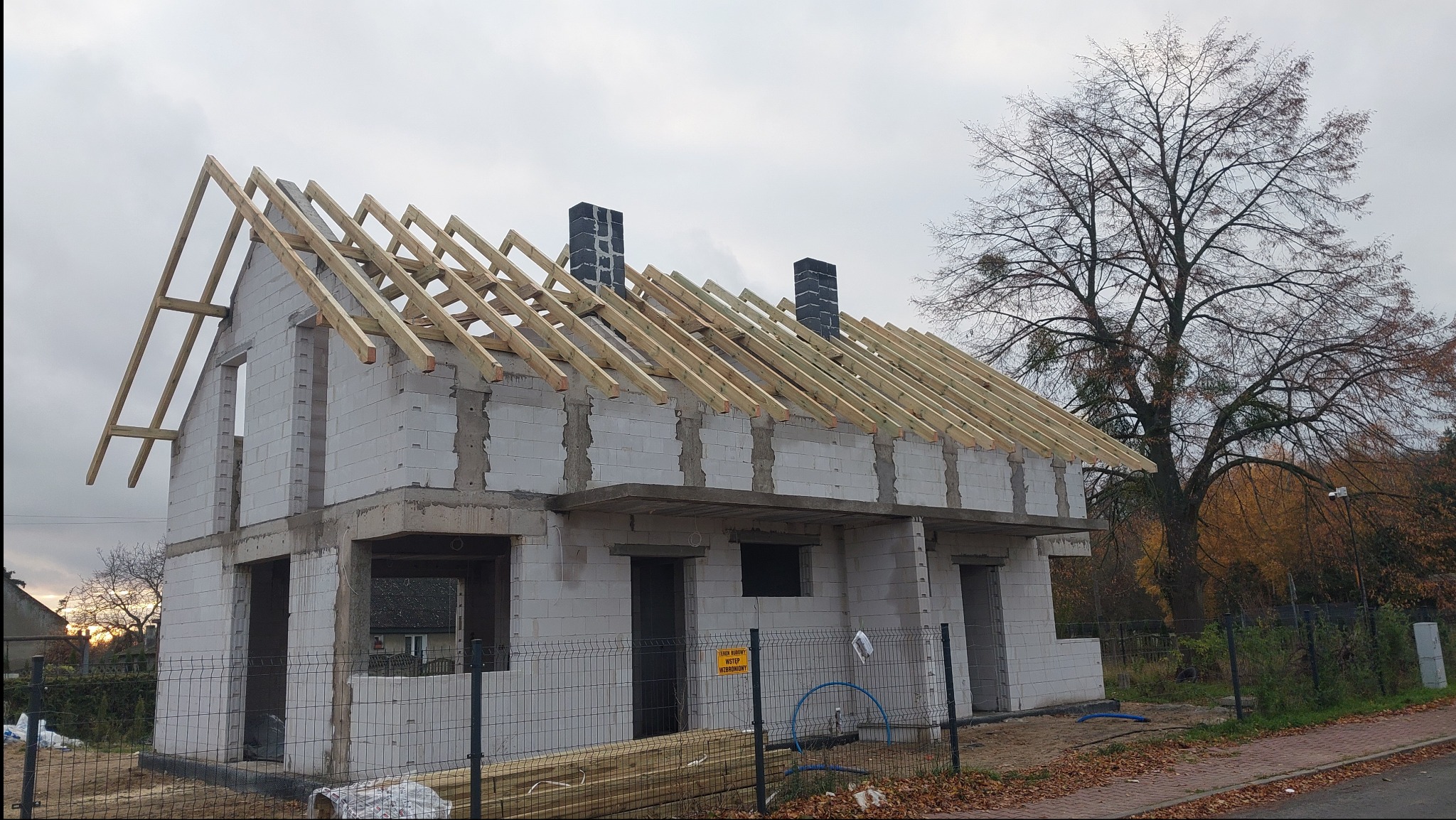 Budowa domu jednorodzinnego w trakcie realizacji. Widoczna surowa konstrukcja ścian z bloczków i drewniana więźba dachowa. Na pierwszym planie tymczasowe ogrodzenie.