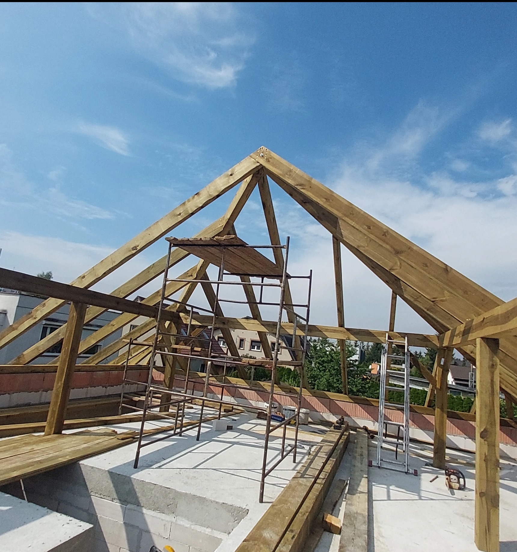 Wznoszenie drewnianej więźby dachowej na murowanym budynku, widok z góry na konstrukcję z rusztowaniem i drabiną na tle błękitnego nieba.