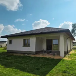 Parterowy dom z ciemnym dachem i jasną elewacją w słoneczny dzień. Widoczne okna z drewnianymi ramami i przesuwne drzwi tarasowe.