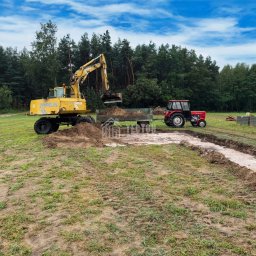 Tecton - Żółta koparka Zeppelin nabiera ziemię do przyczepy ciągniętej przez czerwony traktor na działce z wykopanym fundamentem pod budowę domu.