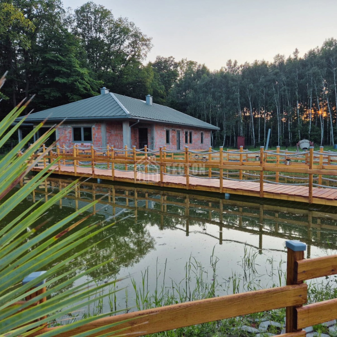 Niedokończony dom z cegły z metalowym dachem, otoczony drewnianym mostem nad stawem, w tle las. Widoczna betoniarka.