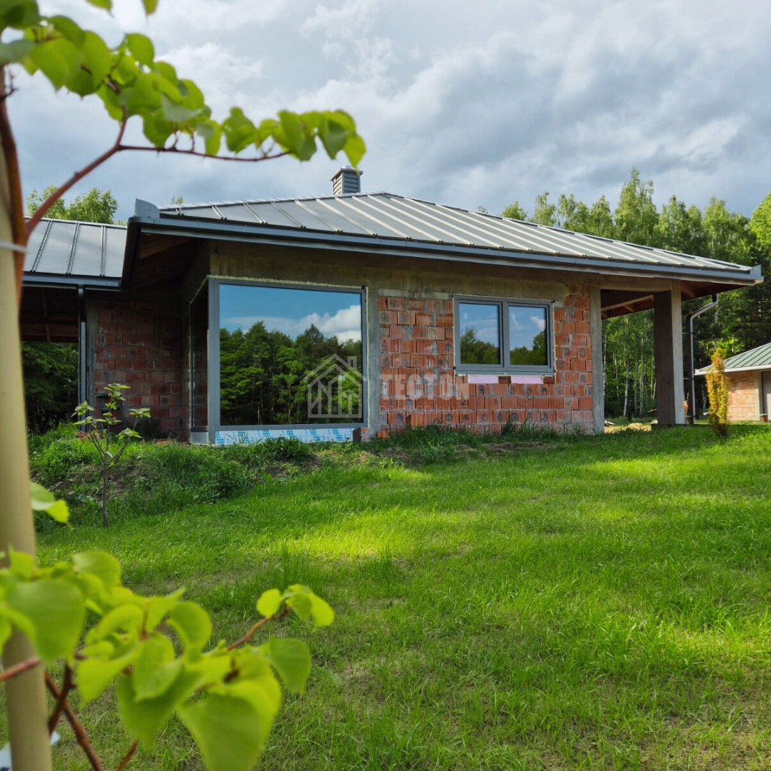Surowy dom z czerwonej cegły z szarym dachem, widoczny przez zielone liście, duże okno odzwierciedla las, budowa w toku, otoczony zielonym trawnikiem.