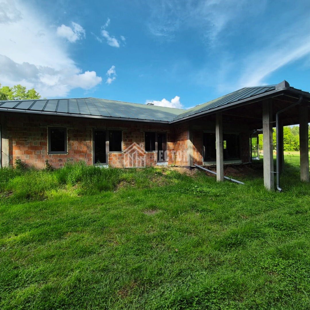Niedokończony dom z czerwonej cegły z ciemnym dachem na zewnątrz, otoczony bujną zielenią, w słoneczny dzień z błękitnym niebem.
