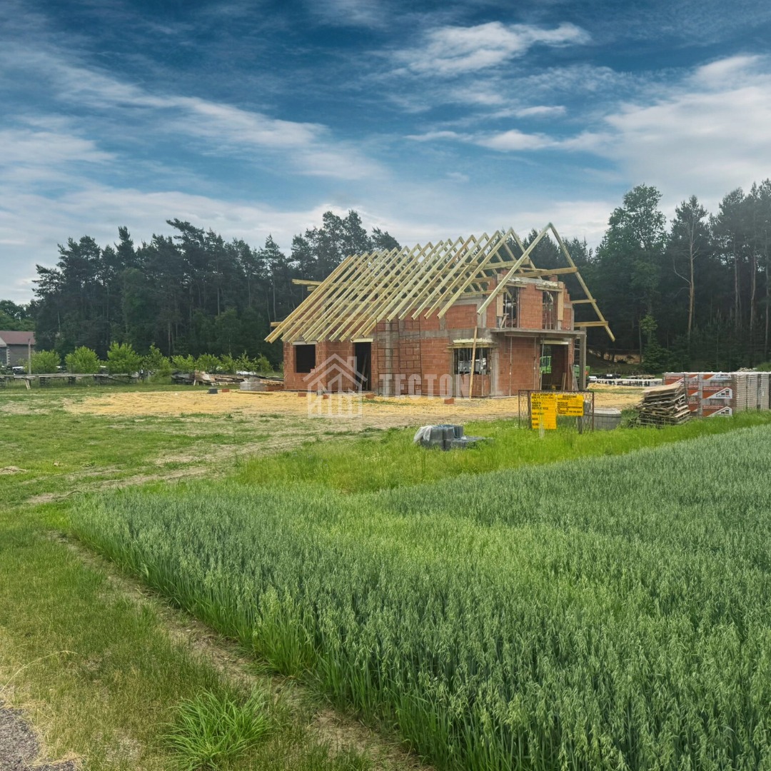 Murowany dom w trakcie budowy, widoczne ściany z czerwonej cegły i drewniana konstrukcja dachu na tle zieleni i lasu. Tablica informacyjna na placu budowy.