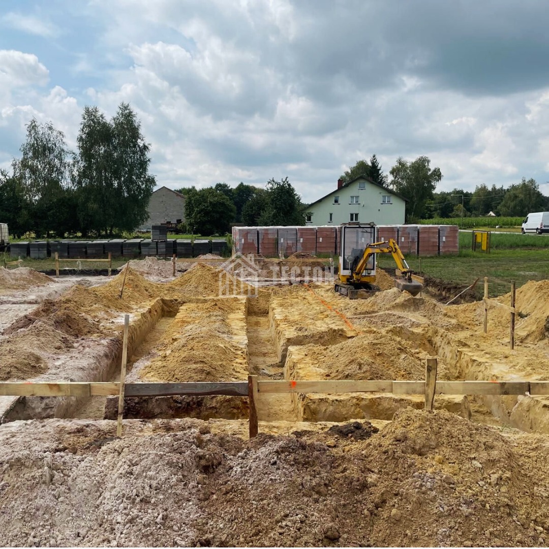 Wykop pod fundamenty domu z widocznym mini-koparką, drewnianym szalunkiem i ułożonymi bloczkami. W tle dom i zieleń. Dzień słoneczny, lekko pochmurny.