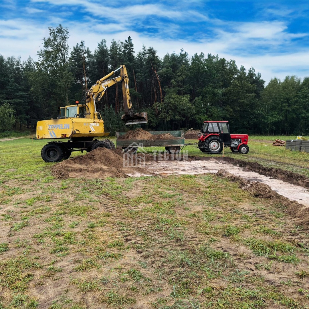 Żółta koparka Zeppelin nabiera ziemię do przyczepy ciągniętej przez czerwony traktor na działce z wykopanym fundamentem pod budowę domu.