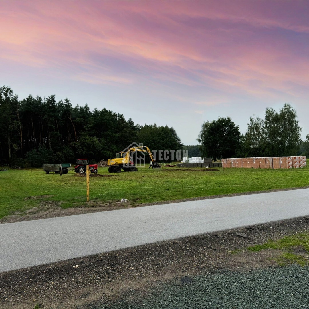 Teren budowy domu z koparką, traktorem i stertą bloczków betonowych na tle lasu i różowego nieba o zmierzchu. Widok z drogi.
