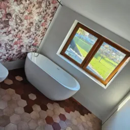 Nowoczesna łazienka z wolnostojącą wanną, heksagonalną podłogą w odcieniach brązu i ścianą z motywem kwiatowym, duże okno z widokiem na zieleń.