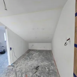 Pokój po szpachlowaniu: białe ściany, szpachla na podłodze, narzędzie. Widoczne kable, gniazdko i fragment drzwi. Skos na suficie. Prace wykończeniowe.