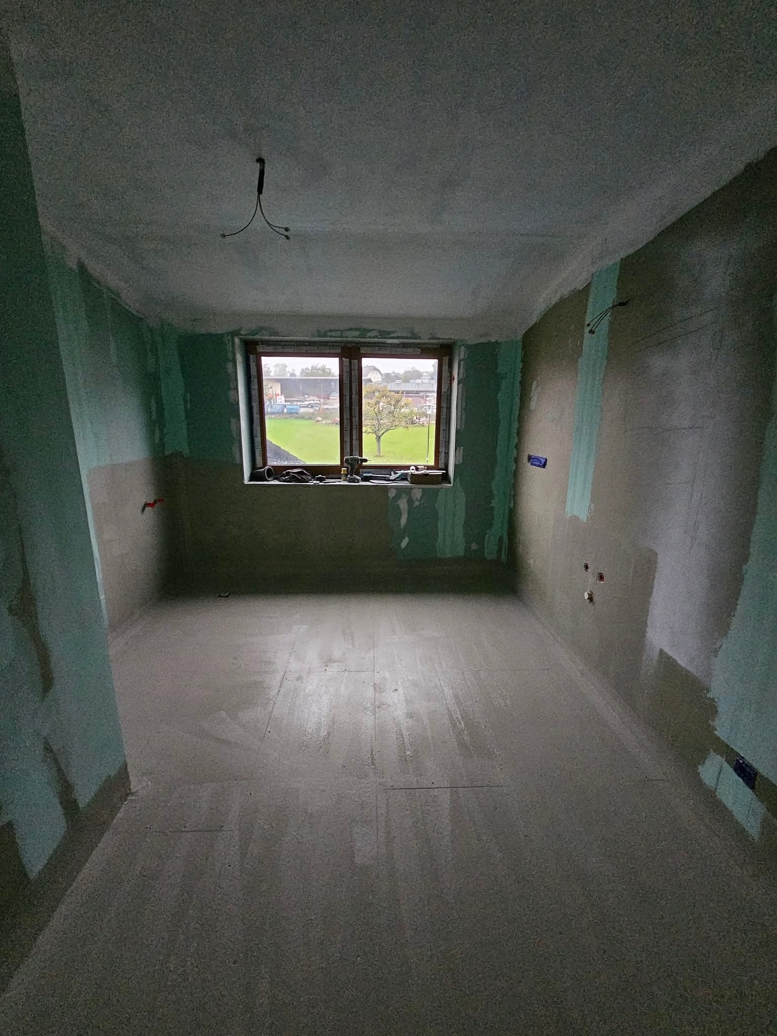 Wnętrze pokoju w trakcie prac wykończeniowych. Widoczne ściany częściowo pokryte gładzią, okno z narzędziami i elementami instalacji elektrycznej. Surowy stan wykończenia.