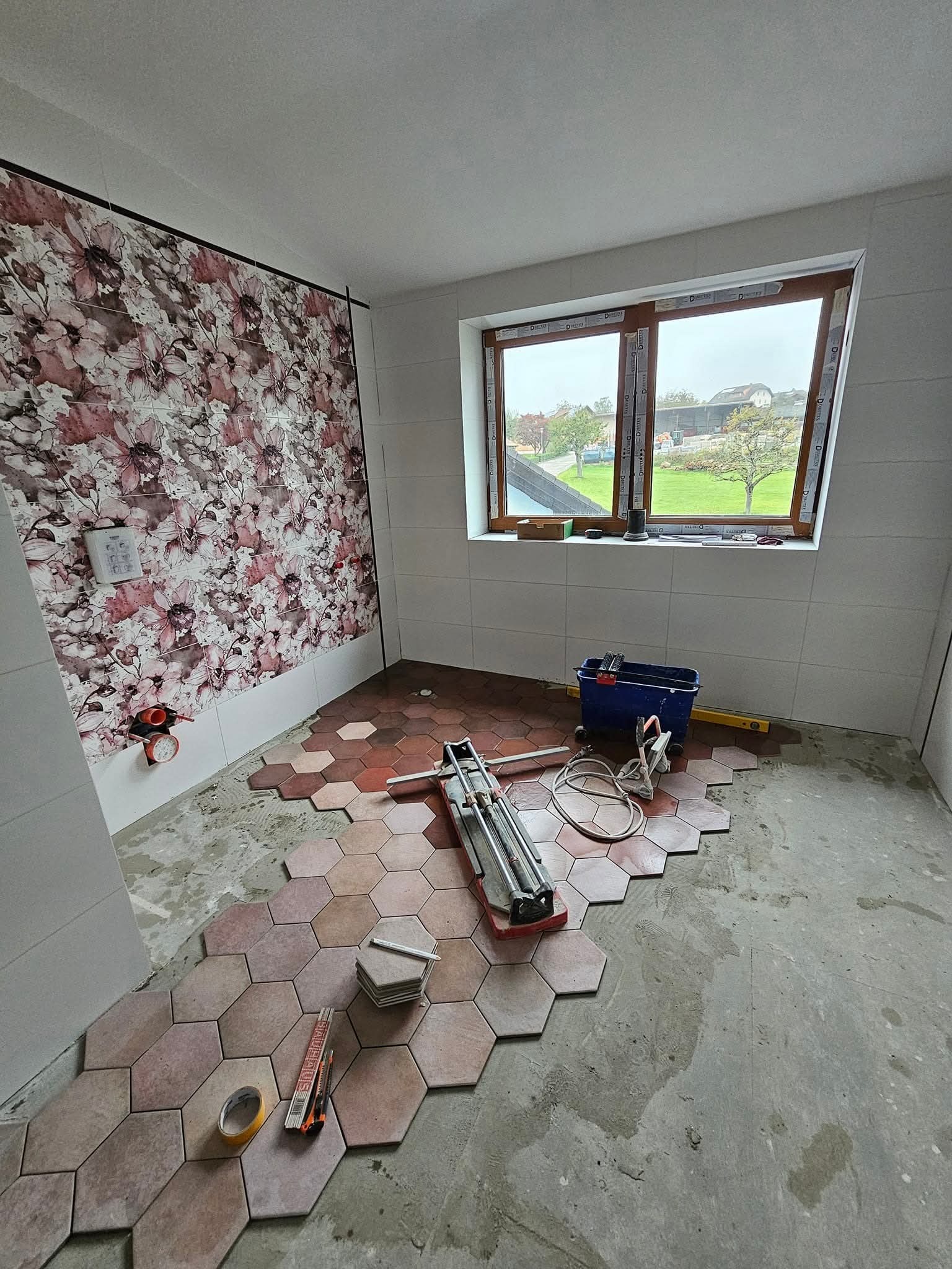 Układanie heksagonalnych płytek w łazience, widoczny fragment ściany z kwiatowym motywem, narzędzia glazurnicze i okno z widokiem na zieleń.