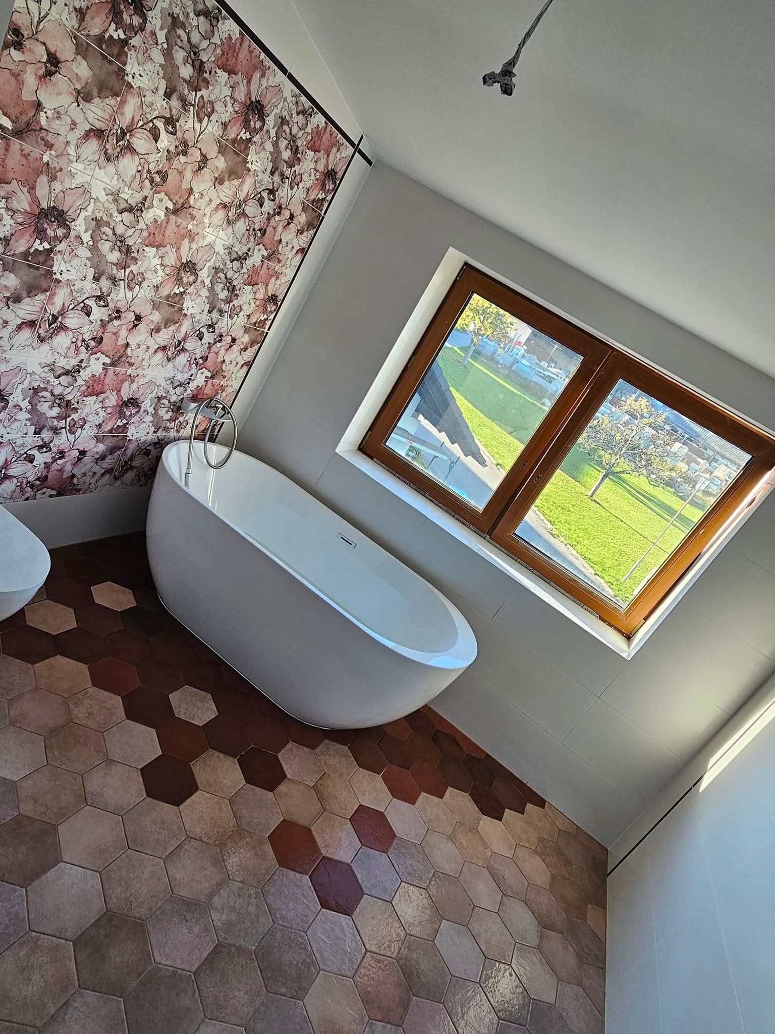 Nowoczesna łazienka z wolnostojącą wanną, heksagonalną podłogą w odcieniach brązu i ścianą z motywem kwiatowym, duże okno z widokiem na zieleń.
