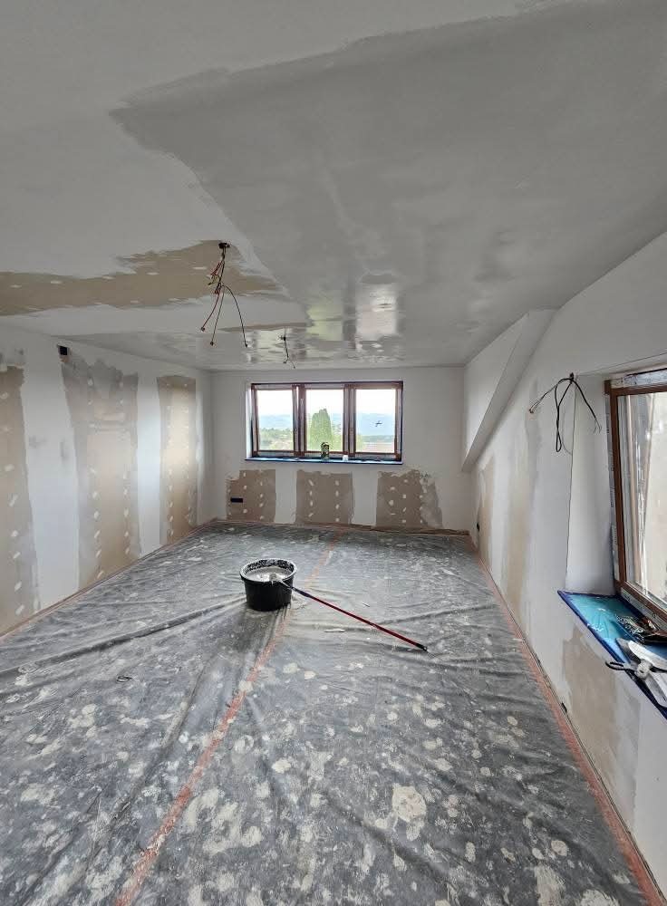 Pokój w trakcie szpachlowania: zabezpieczona folią podłoga, częściowo wyszpachlowane ściany i sufit, widoczne przewody elektryczne, wiadro z zaprawą.