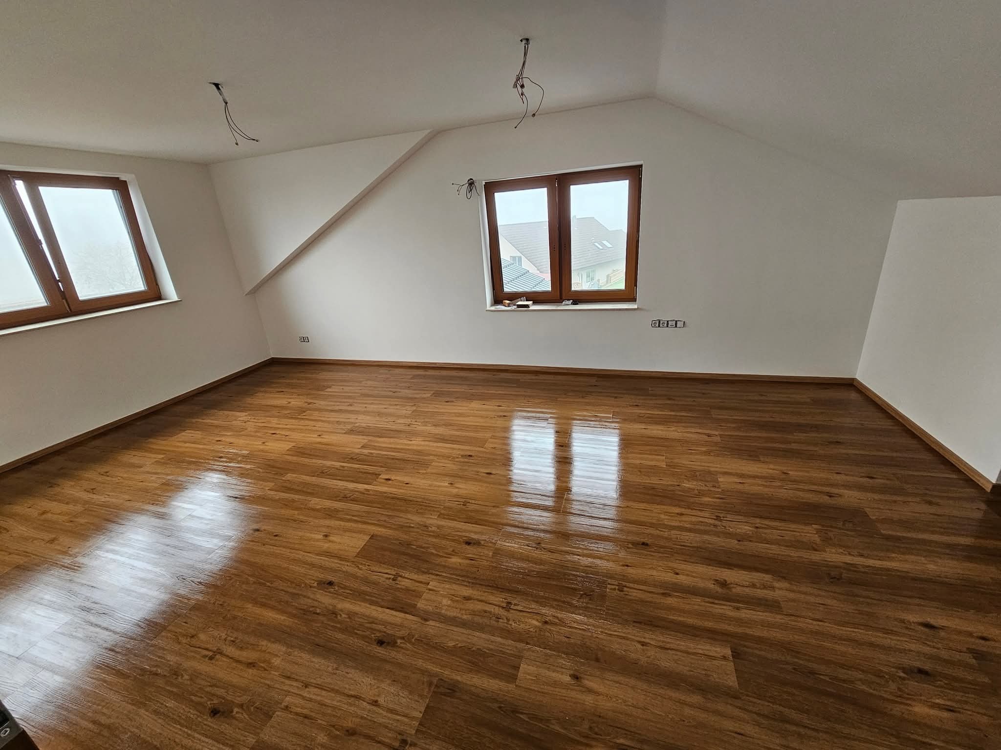 Jasne poddasze w Toruniu po remoncie: świeżo położona, błyszcząca podłoga drewniana, białe ściany, okna z drewnianymi ramami i wystające kable z sufitu.