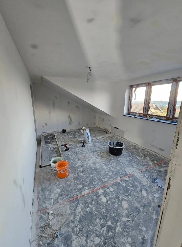 Pokój na poddaszu w trakcie remontu: przygotowanie do malowania ścian, widoczne narzędzia, wiaderka z farbą i gładzią, okno z widokiem na zewnątrz.