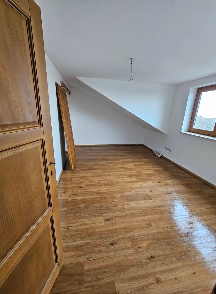 Jasne drewniane panele podłogowe w pokoju na poddaszu z oknem i skośnym sufitem. Widoczne otwarte drewniane drzwi z ozdobnymi panelami.