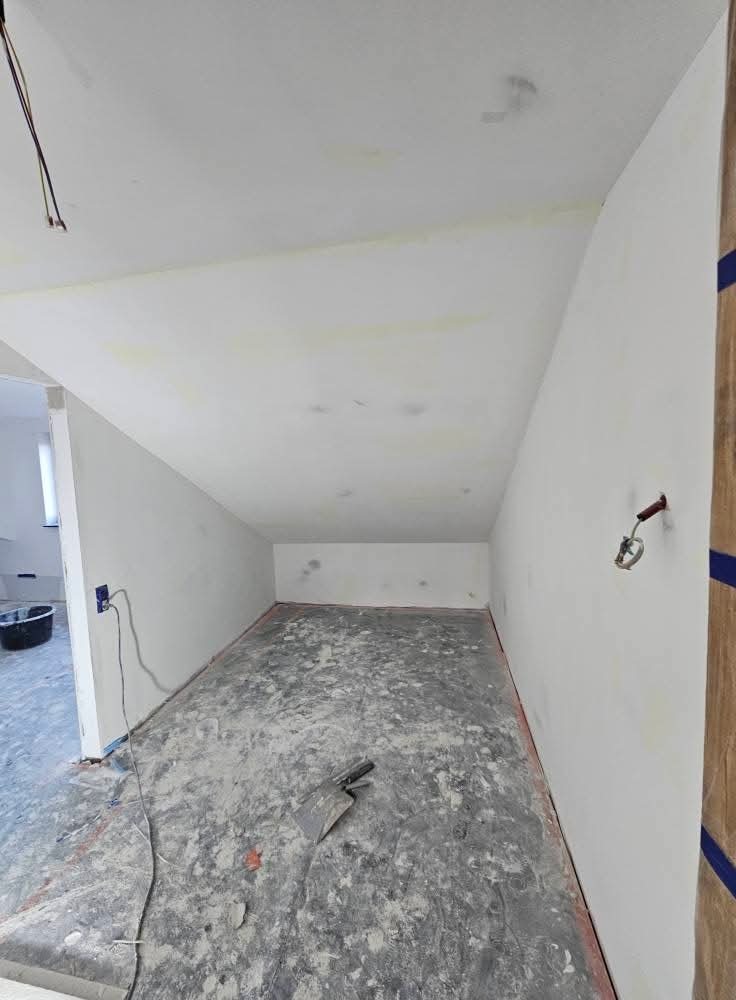 Pokój po szpachlowaniu: białe ściany, szpachla na podłodze, narzędzie. Widoczne kable, gniazdko i fragment drzwi. Skos na suficie. Prace wykończeniowe.