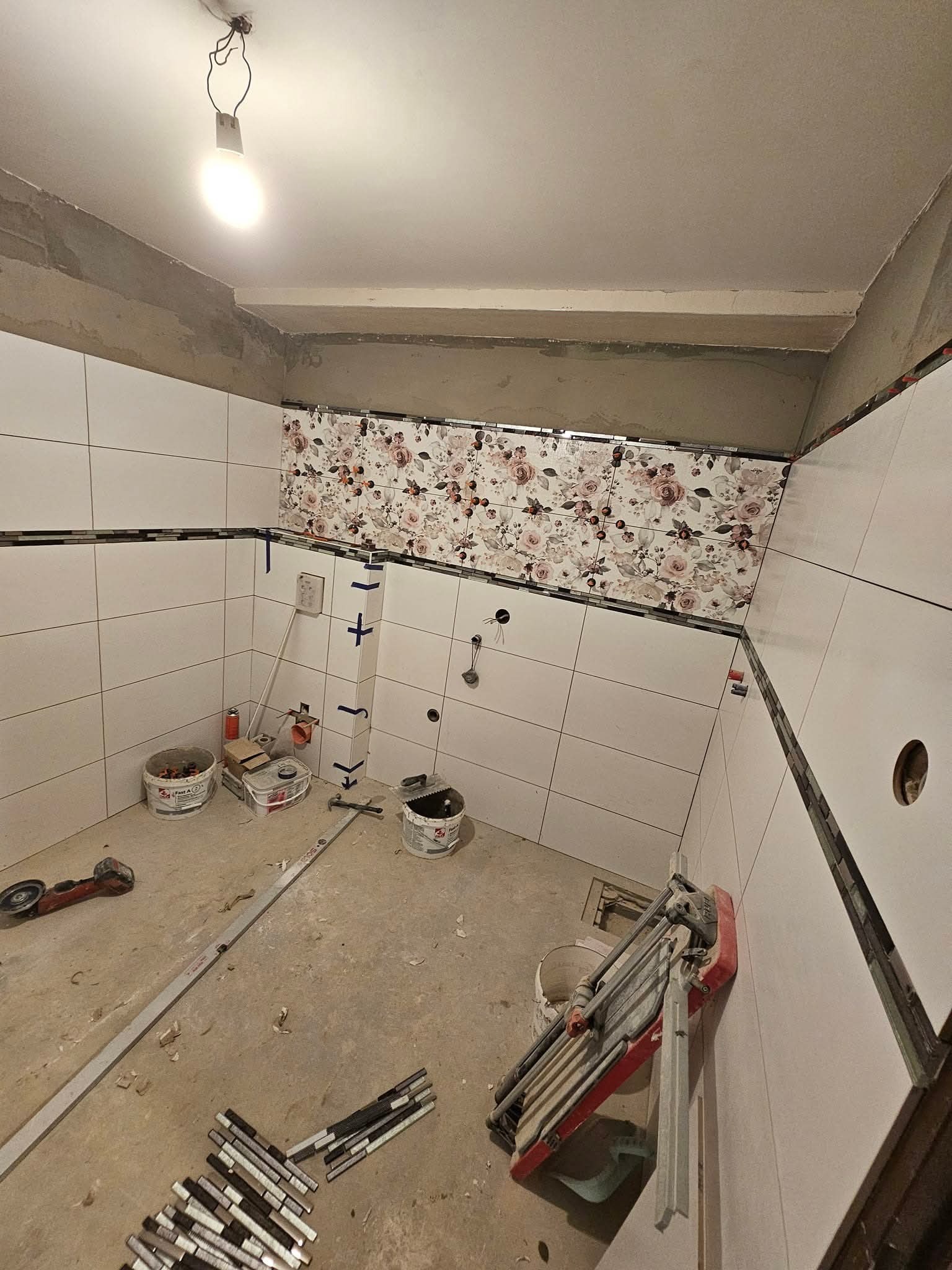 Wnętrze łazienki w trakcie prac glazurniczych: białe płytki na ścianach, dekoracyjny pas z motywem kwiatowym, narzędzia glazurnicze na podłodze, surowy stan wykończenia.