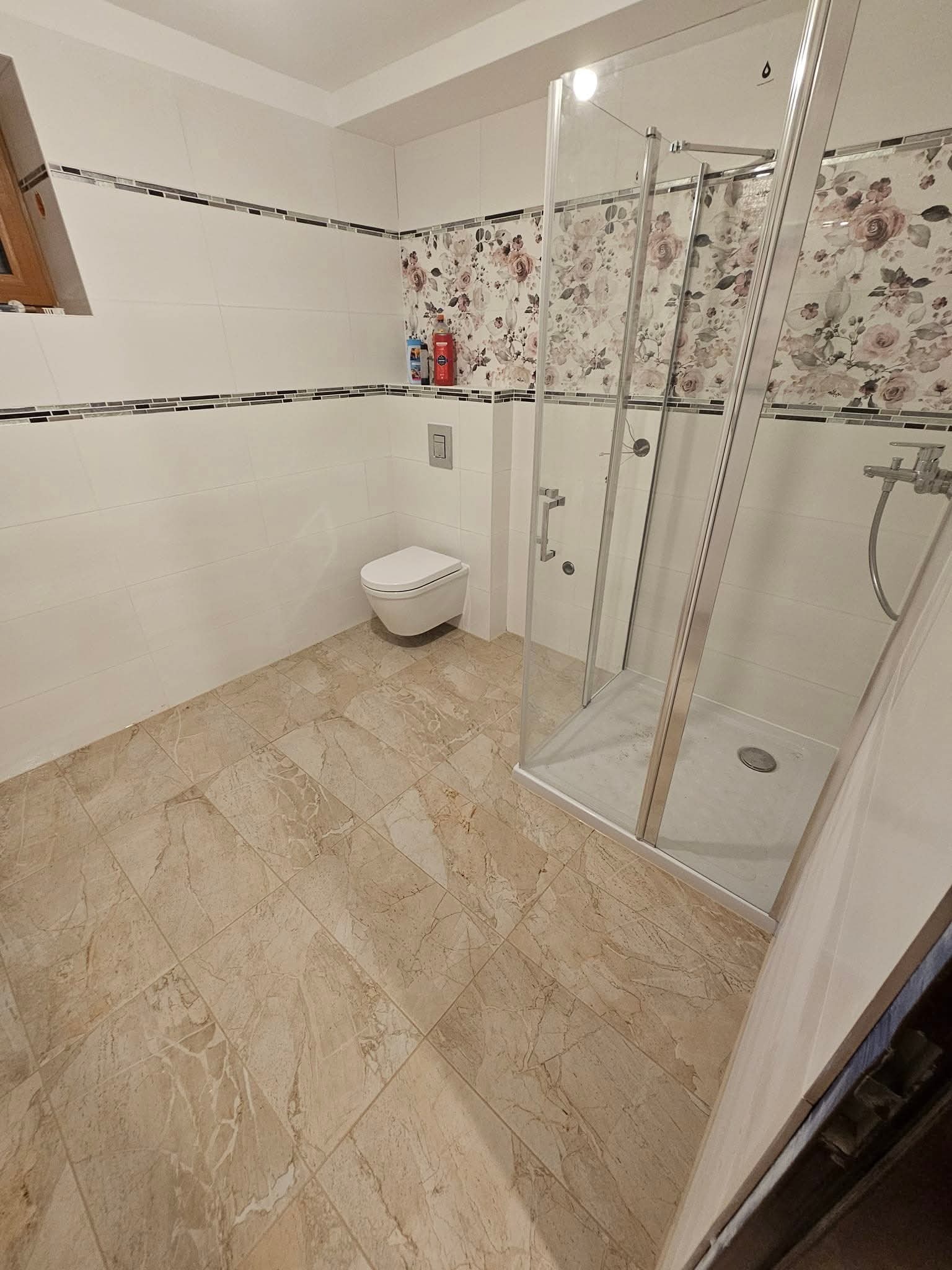 Wykończona łazienka z kabiną prysznicową, beżowymi płytkami podłogowymi i białymi płytkami ściennymi z dekoracyjnym pasem mozaiki i kwiatowym akcentem.