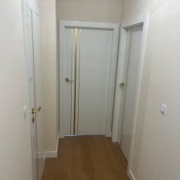 sunrise - Korytarz z trzema białymi drzwiami wewnętrznymi z złotymi akcentami i drewnianą podłogą. Widok na wprost, neutralne oświetlenie.