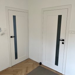 sunrise - Dwa białe, nowoczesne drzwi wewnętrzne z czarnymi klamkami i szklanymi panelami, zamontowane w białym pomieszczeniu z podłogą w jodełkę i szarym dywanikiem.