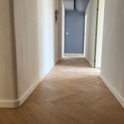 sunrise - Jasny parkiet ułożony w jodełkę w korytarzu z kremowymi ścianami i niebieskim sufitem. Widoczne drzwi po prawej stronie z zieloną ościeżnicą i niebieskie drzwi na końcu korytarza.