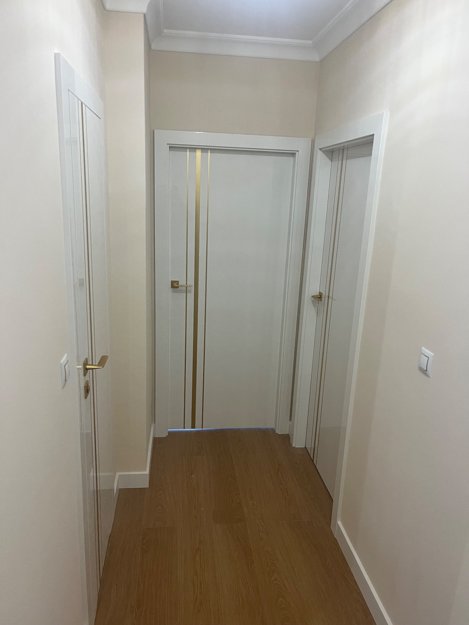 Korytarz z trzema białymi drzwiami wewnętrznymi z złotymi akcentami i drewnianą podłogą. Widok na wprost, neutralne oświetlenie.