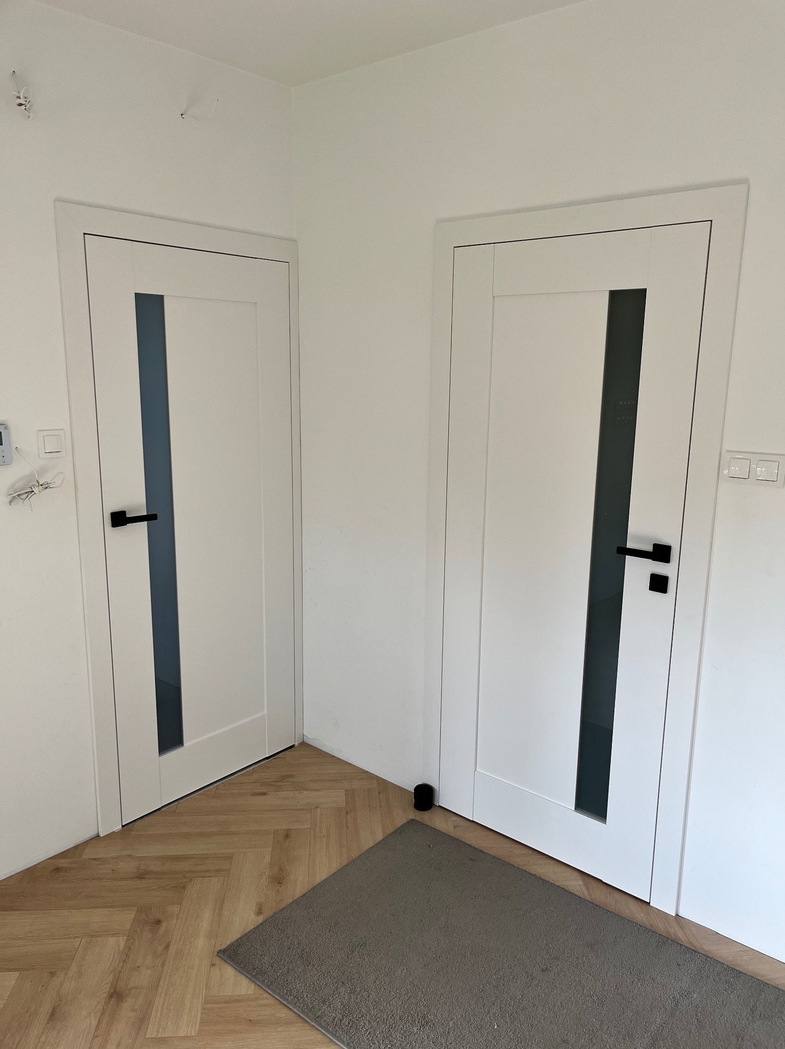 Dwa białe, nowoczesne drzwi wewnętrzne z czarnymi klamkami i szklanymi panelami, zamontowane w białym pomieszczeniu z podłogą w jodełkę i szarym dywanikiem.