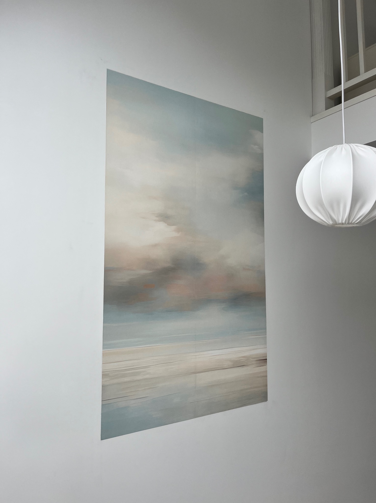 Abstrakcyjny obraz z motywem pejzażu morskiego zawieszony na białej ścianie, obok lampa w kształcie kuli i fragment okna. Minimalistyczny wystrój wnętrza.