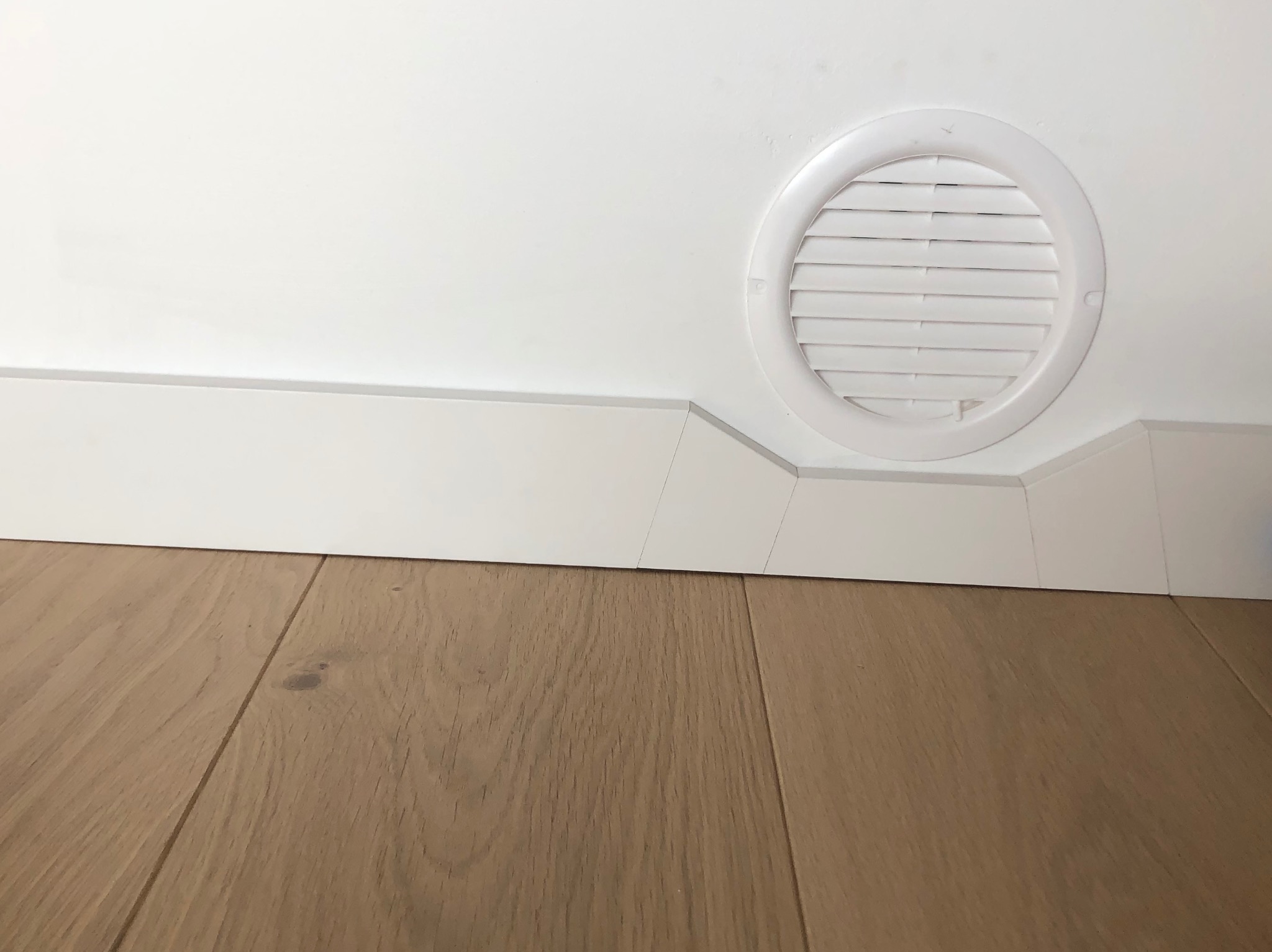 Jasna drewniana podłoga z listwą przypodłogową i okrągłą kratką wentylacyjną w białej ścianie. Minimalistyczny detal wykończenia wnętrza.