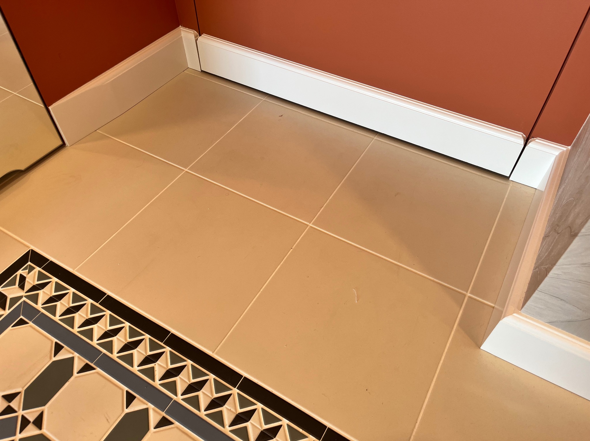 Beżowe płytki podłogowe z dekoracyjnym, geometrycznym wzorem przy ścianie w kolorze terakoty z białą listwą przypodłogową. Widoczny fragment lustra.