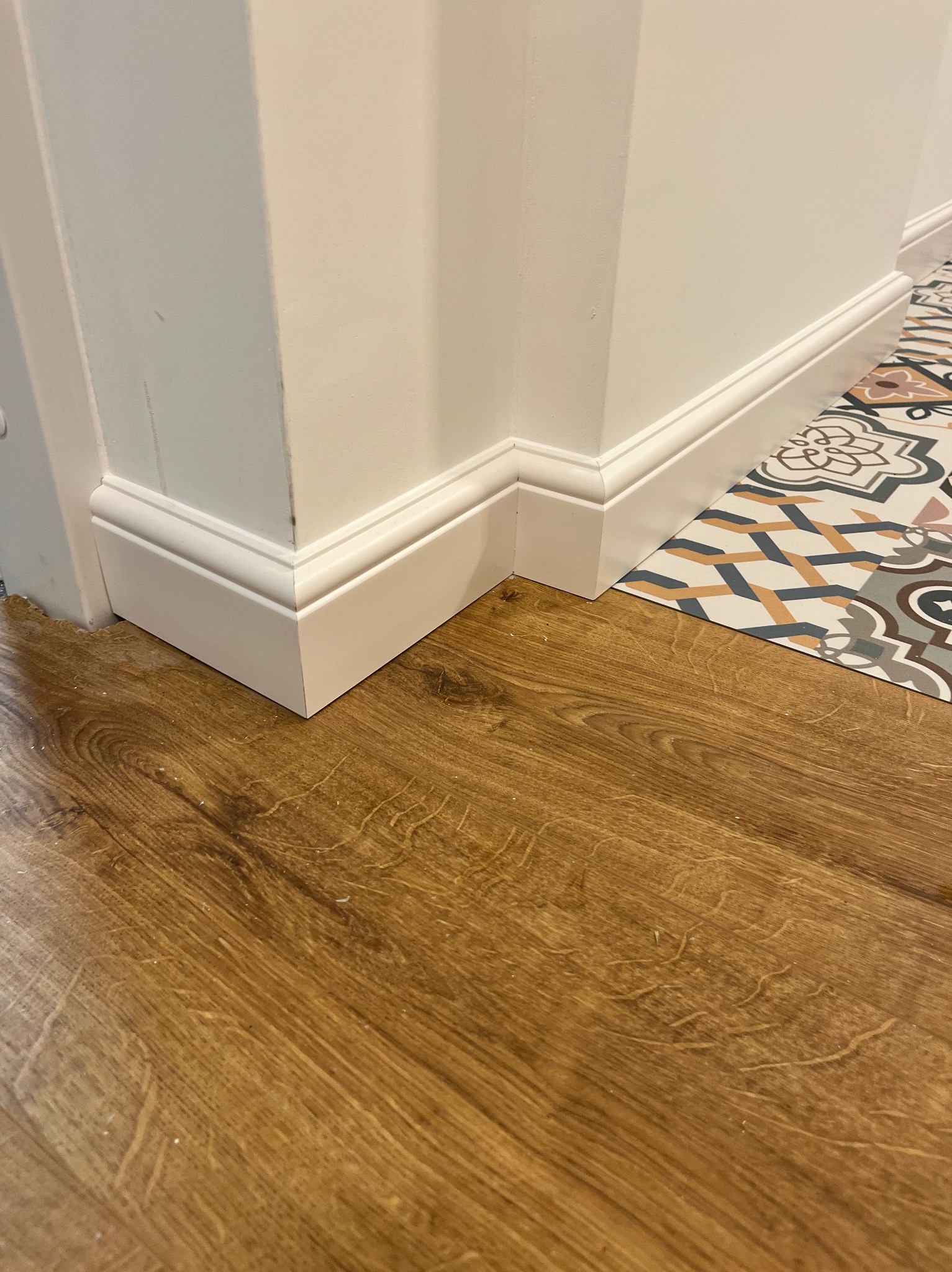 Parkiet w odcieniach brązu, biała listwa przypodłogowa przy ścianie i fragment wzorzystej podłogi w rogu, ukazujące połączenie różnych stylów wykończenia.