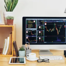 Monitor z wykresami giełdowymi na biurku z roślinami, tabletem i akcesoriami, oświetlony lampką. Układ biurka sugeruje pracę analityczną.