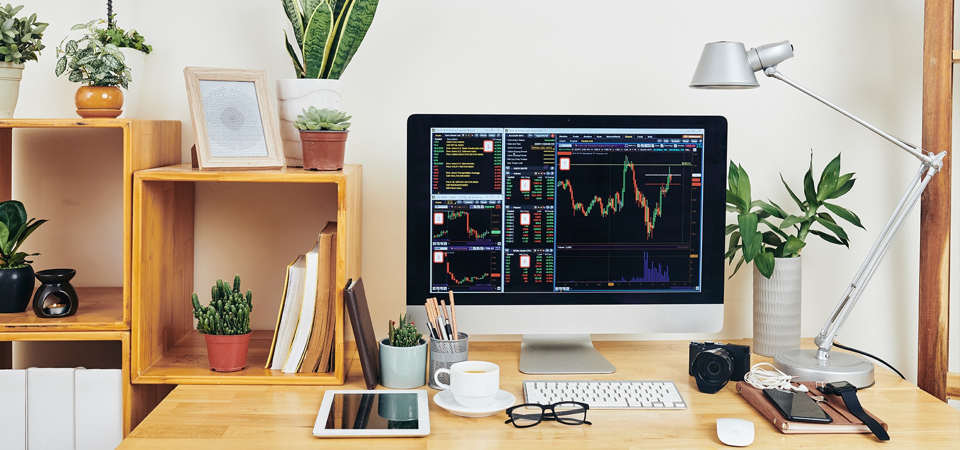 Monitor z wykresami giełdowymi na biurku z roślinami, tabletem i akcesoriami, oświetlony lampką. Układ biurka sugeruje pracę analityczną.