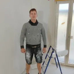 Mężczyzna w ubraniach roboczych stoi w rogu pomalowanego na biało pokoju, obok drabiny. Ściany świeżo pomalowane, okno zabezpieczone folią malarską.