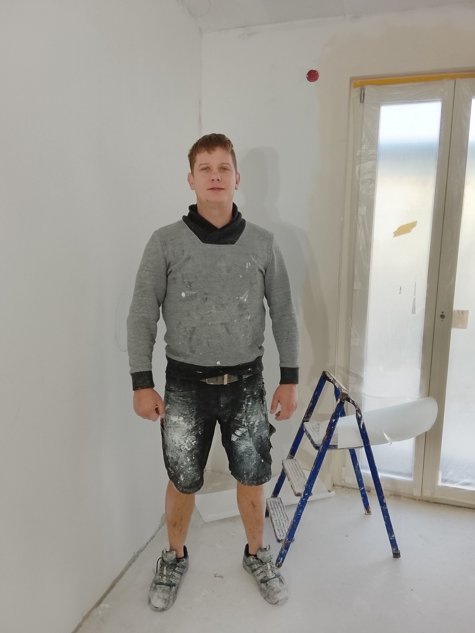 Mężczyzna w ubraniach roboczych stoi w rogu pomalowanego na biało pokoju, obok drabiny. Ściany świeżo pomalowane, okno zabezpieczone folią malarską.