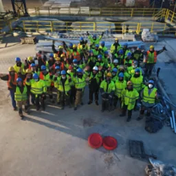 Zdjęcie grupy pracowników w kaskach i kamizelkach odblaskowych, stojących na placu budowy i machających do kamery. W tle widoczne konstrukcje i elementy infrastruktury.