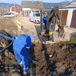 ZBART ELECTRIC Krzysztof Frysztak - Prace ziemne: operator koparki i pracownik z łopatą przy wykopie z niebieskimi rurami. W tle budynki i pojazdy na tle błękitnego nieba.