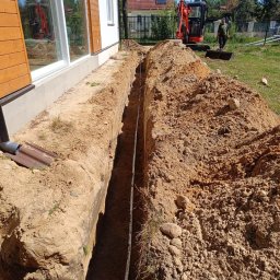 ZBART ELECTRIC Krzysztof Frysztak - Wykopany rów wzdłuż domu z widoczną rurą, minikoparka i pracownik w tle. Przygotowanie do instalacji podziemnej.