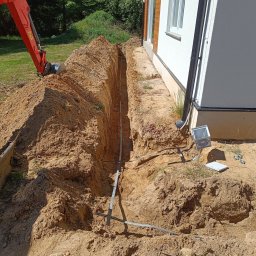 ZBART ELECTRIC Krzysztof Frysztak - Wykop pod instalację elektryczną przy białym budynku jednorodzinnym. W wykopie widoczne rury i przewody. W tle koparka.