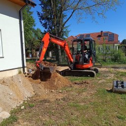 ZBART ELECTRIC Krzysztof Frysztak - Pomarańczowa minikoparka Kubota w trakcie prac ziemnych przy budynku jednorodzinnym. Widoczny operator i wykopany grunt. W tle dom z panelami słonecznymi.